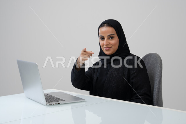 Saudi female office job and profession, managing and completing tasks using a modern and advanced technical device, portrait of a Saudi Arabian Gulf woman wearing a hijab and black abaya pointing to the camera working on a laptop at the company headquarters, gray background