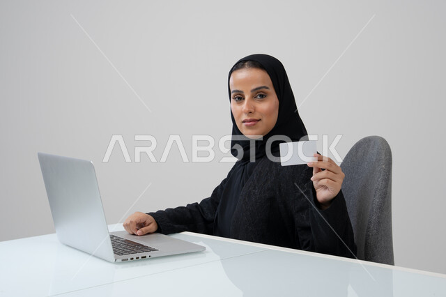 عرض موك اب ابيض صغير، وظيفة ومهنة نسائية مكتبية سعودية، ادارة وانجاز المهام باستخدام جهاز تقني حديث ومتطور, بورتريه لإمرأة عربية خليجية سعودية مبتسمة ترتدي الحجاب والعباءة السوداء تمسك بيدها بطاقة تعريفية تعمل على الحاسب المحمول, خلفية رمادية