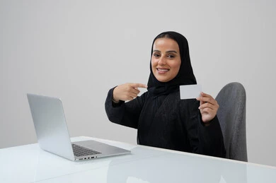 عرض موك اب ابيض صغير، وظيفة ومهنة نسائية مكتبية سعودية، ادارة وانجاز المهام باستخدام جهاز تقني حديث ومتطور, بورتريه لإمرأة عربية خليجية سعودية مبتسمة ترتدي الحجاب والعباءة السوداء تمسك بيدها بطاقة تعريفية تشير اليها تعمل على الحاسب المحمول, خلفية رمادية