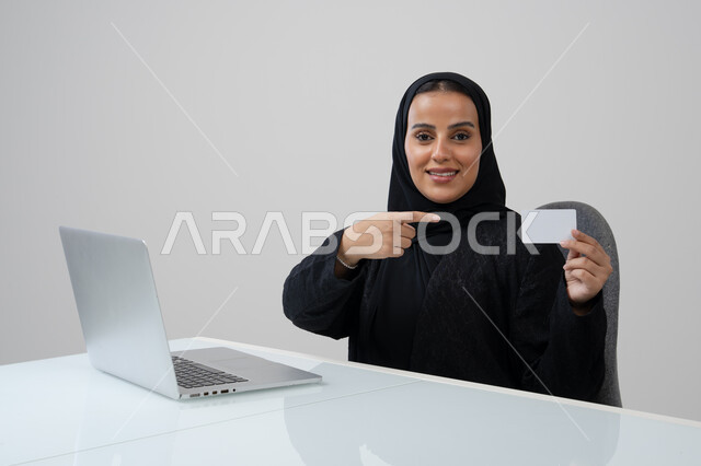 عرض موك اب ابيض صغير، وظيفة ومهنة نسائية مكتبية سعودية، ادارة وانجاز المهام باستخدام جهاز تقني حديث ومتطور, بورتريه لإمرأة عربية خليجية سعودية مبتسمة ترتدي الحجاب والعباءة السوداء تمسك بيدها بطاقة تعريفية تشير اليها تعمل على الحاسب المحمول, خلفية رمادية