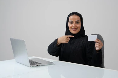 Small white mockup, Saudi female office job and profession, managing and completing tasks using a modern and advanced technical device, portrait of a smiling Saudi Arabian Gulf woman wearing a hijab and black abaya holding an ID card in her hand pointing to it working on a laptop, gray background
