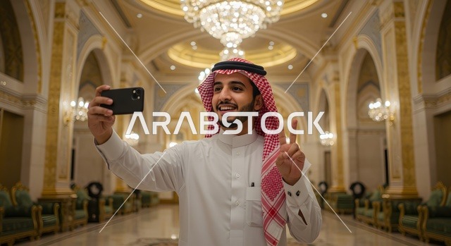 الاحتفاظ بالصور واللحظات التذكارية، رجل عربي خليجي سعودي مبتسم يرتدي الثوب والشماغ التقليدي يقوم بالتقاط صورة سيلفي باستخدام الهاتف المحمول في قاعة فاخرة، استخدام التقنية الحديقة والمطورة، ايماءات السعادة والسرور