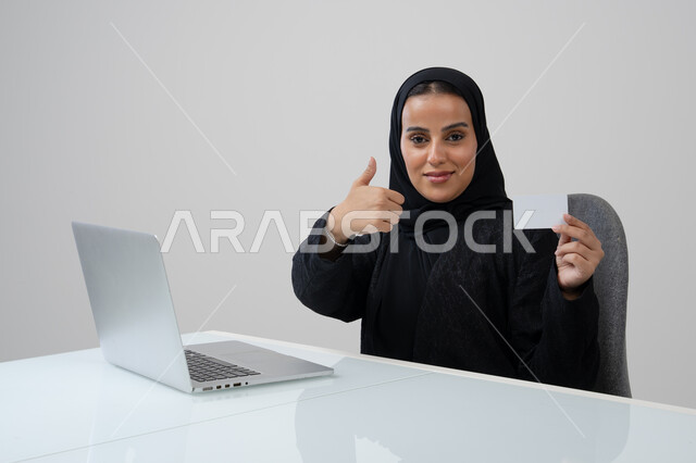 رفع اصبع الابهام للأعلى بتعابير الرضا والاعجاب، عرض موك اب ابيض، وظيفة نسائية مكتبية، ادارة وانجاز المهام باستخدام الجهاز التقني, بورتريه لإمرأة عربية خليجية سعودية ترتدي الحجاب والعباءة تمسك بيدها بطاقة تعريفية تشير اليها تعمل على الحاسب المحمول, خلفية رمادية