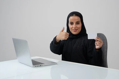 رفع اصبع الابهام للأعلى بتعابير الرضا والاعجاب، عرض موك اب ابيض، وظيفة نسائية مكتبية، ادارة وانجاز المهام باستخدام الجهاز التقني, بورتريه لإمرأة عربية خليجية سعودية ترتدي الحجاب والعباءة تمسك بيدها بطاقة تعريفية تشير اليها تعمل على الحاسب المحمول, خلفية رمادية