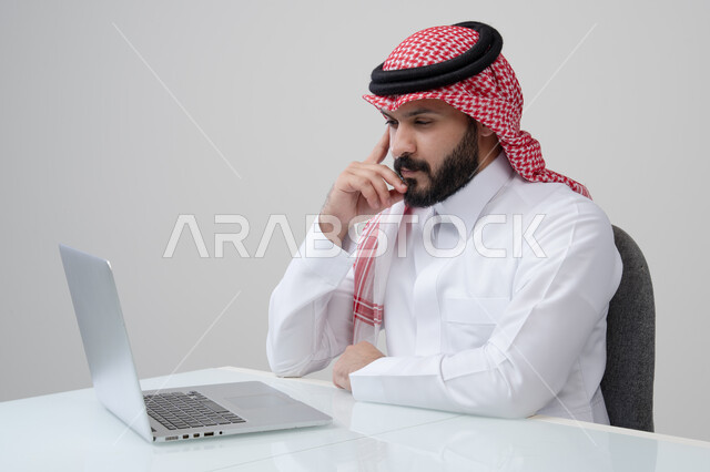 ايجاد حل لمشكلة ما، مهنة ووظيفة ادارية، استخدام التقنية الحديثه المتطورة في الحياة العملية، ايماءات التفكير والتركيز، بورتريه لرجل عربي خليجي سعودي يرتدي الثوب التقليدي والشماغ يضع يده على خده بتعابير شرود الذهن، العمل على الحاسب المحمول، خلفية رمادية