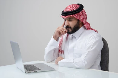 ايجاد حل لمشكلة ما، مهنة ووظيفة ادارية، استخدام التقنية الحديثه المتطورة في الحياة العملية، ايماءات التفكير والتركيز، بورتريه لرجل عربي خليجي سعودي يرتدي الثوب التقليدي والشماغ يضع يده على خده بتعابير شرود الذهن، العمل على الحاسب المحمول، خلفية رمادية
