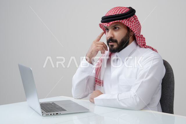 ايجاد حل لمشكلة ما، مهنة ووظيفة ادارية، استخدام التقنية الحديثه المتطورة في الحياة العملية، ايماءات التفكير والتركيز، بورتريه لرجل عربي خليجي سعودي يرتدي الثوب التقليدي والشماغ يضع يده على خده بتعابير شرود الذهن، العمل على الحاسب المحمول، خلفية رمادية