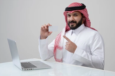 عرض موك اب ابيض، مهنة ووظيفة ادارية، استخدام التقنية الحديثه المتطورة في الحياة العملية، بورتريه لرجل عربي خليجي سعودي مبتسم يرتدي الثوب التقليدي والشماغ يمسك بيده بطاقة تعريفية يشير اليها، العمل على الحاسب المحمول في مقر الشركة، خلفية رمادية