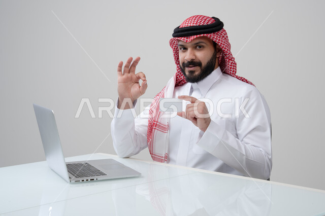 عرض موك اب ابيض، مهنة ووظيفة ادارية، استخدام التقنية الحديثه في الحياة العملية، بورتريه لرجل عربي خليجي سعودي مبتسم يرتدي الثوب التقليدي والشماغ يمسك بيده بطاقة تعريفية يرفع يده لأعلى بتعابير الرضا والقبول والاعجاب، العمل على الحاسب المحمول، خلفية رمادية