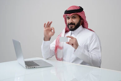 عرض موك اب ابيض، مهنة ووظيفة ادارية، استخدام التقنية الحديثه في الحياة العملية، بورتريه لرجل عربي خليجي سعودي مبتسم يرتدي الثوب التقليدي والشماغ يمسك بيده بطاقة تعريفية يرفع يده لأعلى بتعابير الرضا والقبول والاعجاب، العمل على الحاسب المحمول، خلفية رمادية
