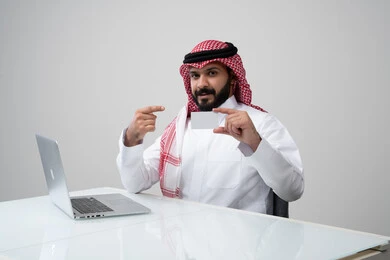 عرض موك اب ابيض، مهنة ووظيفة ادارية، استخدام التقنية الحديثه المتطورة في الحياة العملية، بورتريه لرجل عربي خليجي سعودي مبتسم يرتدي الثوب التقليدي والشماغ يمسك بيده بطاقة تعريفية يشير اليها، العمل على الحاسب المحمول في مقر الشركة، خلفية رمادية
