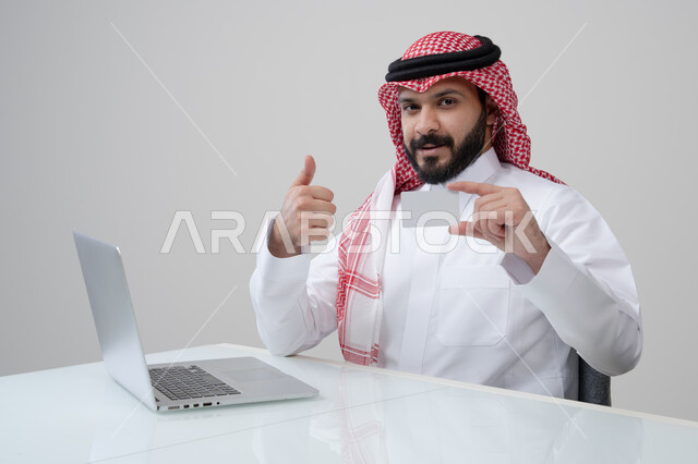 عرض موك اب ابيض، مهنة ووظيفة ادارية، استخدام التقنية الحديثه في الحياة العملية، بورتريه لرجل عربي خليجي سعودي مبتسم يرتدي الثوب التقليدي والشماغ يمسك بيده بطاقة تعريفية يرفع اصبع ابهام بتعابير الرضا والقبول والاعجاب، العمل على الحاسب المحمول، خلفية رمادية