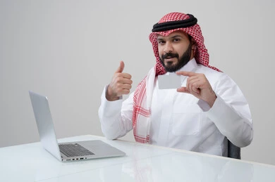 White mockup presentation, administrative profession and job, use of modern technology in practical life, portrait of a smiling Saudi Arabian Gulf man wearing traditional thobe and shemagh holding an ID card and raising a thumbs up with expressions of satisfaction, acceptance and admiration, working on a laptop, gray background