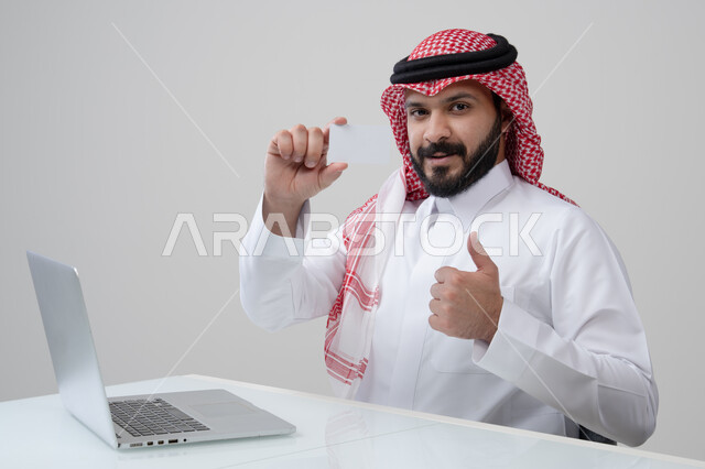 عرض موك اب ابيض، مهنة ووظيفة ادارية، استخدام التقنية الحديثه في الحياة العملية، بورتريه لرجل عربي خليجي سعودي مبتسم يرتدي الثوب التقليدي والشماغ يمسك بيده بطاقة تعريفية يرفع اصبع ابهام بتعابير الرضا والقبول والاعجاب، العمل على الحاسب المحمول، خلفية رمادية