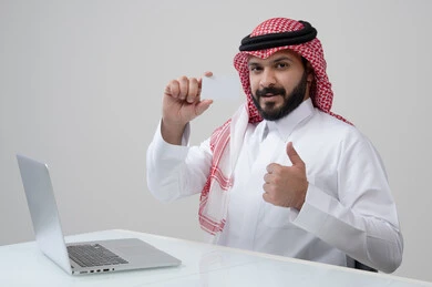 عرض موك اب ابيض، مهنة ووظيفة ادارية، استخدام التقنية الحديثه في الحياة العملية، بورتريه لرجل عربي خليجي سعودي مبتسم يرتدي الثوب التقليدي والشماغ يمسك بيده بطاقة تعريفية يرفع اصبع ابهام بتعابير الرضا والقبول والاعجاب، العمل على الحاسب المحمول، خلفية رمادية