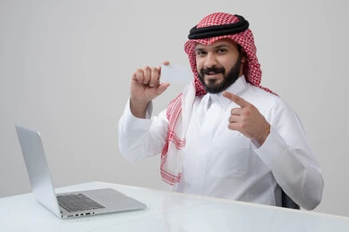 عرض موك اب ابيض، مهنة ووظيفة ادارية، استخدام التقنية الحديثه المتطورة في الحياة العملية، بورتريه لرجل عربي خليجي سعودي مبتسم يرتدي الثوب التقليدي والشماغ يمسك بيده بطاقة تعريفية يشير اليها، العمل على الحاسب المحمول في مقر الشركة، خلفية رمادية