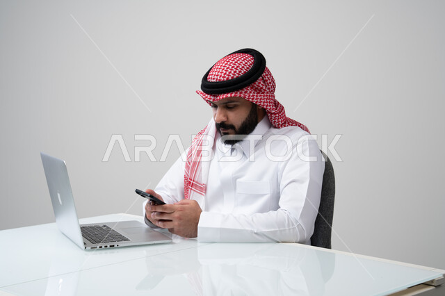 وظيفة ومهنة ادارية مكتبية، استخدام التقنية الحديثه المتطورة في الحياة العملية، بورتريه لرجل عربي خليجي سعودي يرتدي الثوب التقليدي والشماغ يمسك بيده الهاتف المحمول بتعابير الاندماج والتركيز، العمل على الحاسب المحمول، خلفية رمادية