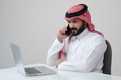 الرد على اتصال هاتفي، وظيفة ادارية مكتبية، استخدام التقنية الحديثه المتطورة في الحياة العملية، بورتريه لرجل عربي خليجي سعودي يرتدي الثوب التقليدي والشماغ يجري مكالمة صوتية عبر الهاتف المحمول بتعابير الاندماج والتركيز، العمل على الحاسب المحمول، خلفية رمادية