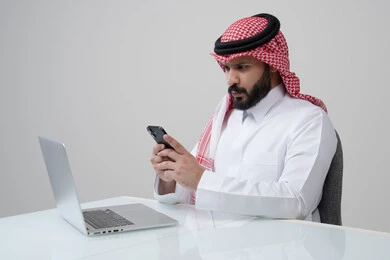 وظيفة ومهنة ادارية مكتبية، استخدام التقنية الحديثه المتطورة في الحياة العملية، بورتريه لرجل عربي خليجي سعودي يرتدي الثوب التقليدي والشماغ يمسك بيده الهاتف المحمول بتعابير الاندماج والتركيز، العمل على الحاسب المحمول، خلفية رمادية