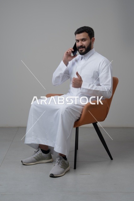 الرد على مكالمة هاتفية عبر الجوال، الدردشة والمحادثة باستخدام جهاز تقني حديث, بورتريه لرجل عربي خليجي سعودي يرتدي الثوب التقليدي يجلس على الكرسي يقوم باجراء اتصال هاتفي من خلال الهاتف المحمول، رفع اصبع الابهام لأعلى بتعابير الاعجاب والقبول، خلفية بيضاء