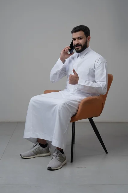 Answering a phone call via mobile phone, chatting and talking using a modern technical device, portrait of a Saudi Arabian Gulf man wearing traditional thobe sitting on a chair making a phone call via mobile phone, raising the thumb up with expressions of admiration and acceptance, white background