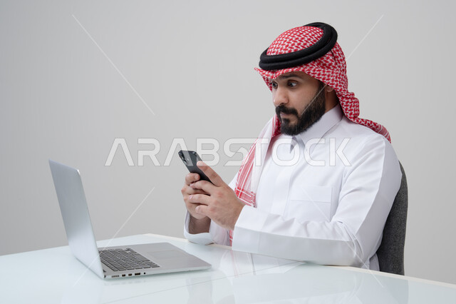 وظيفة ومهنة ادارية مكتبية، استخدام التقنية الحديثه المتطورة في الحياة العملية، بورتريه لرجل عربي خليجي سعودي يرتدي الثوب التقليدي والشماغ يمسك بيده الهاتف المحمول بتعابير الاندماج والتركيز، العمل على الحاسب المحمول، خلفية رمادية