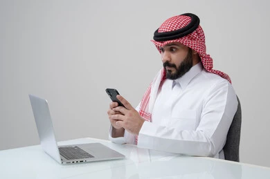 وظيفة ومهنة ادارية مكتبية، استخدام التقنية الحديثه المتطورة في الحياة العملية، بورتريه لرجل عربي خليجي سعودي يرتدي الثوب التقليدي والشماغ يمسك بيده الهاتف المحمول بتعابير الاندماج والتركيز، العمل على الحاسب المحمول، خلفية رمادية