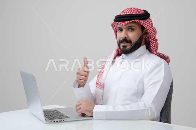 ايماءات الاعجاب والسرور، بورتريه لرجل عربي خليجي سعودي مبتسم يرتدي الثوب التقليدي والشماغ يرفع اصبع الابهام لأعلى بتعابير القبول يقوم بالعمل على الحاسب المحمول، مهنة ووظيفة ادارية، استخدام التقنية الحديثه المتطورة في الحياة العملية، خلفية رمادية