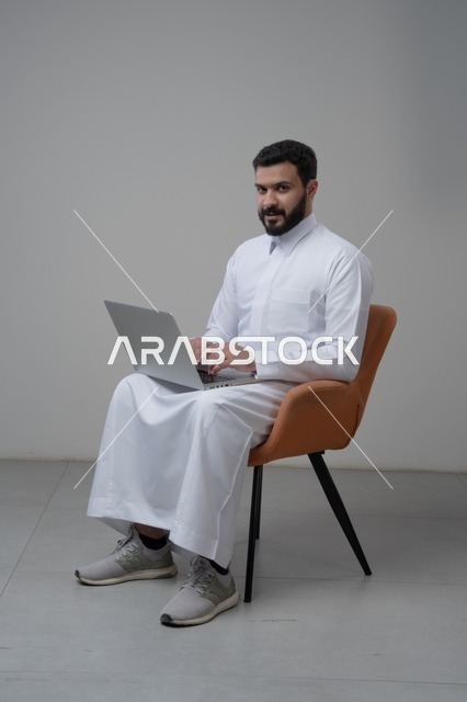 العمل عن بعد، استخدام التقنية الحديثه المتطورة في الحياة العملية، ايماءات الاندماج والتركيز، بورتريه لرجل عربي خليجي سعودي يرتدي الثوب التقليدي والشماغ يقوم بالعمل على الحاسب المحمول يجلس على كرسي مريح، خلفية بيضاء