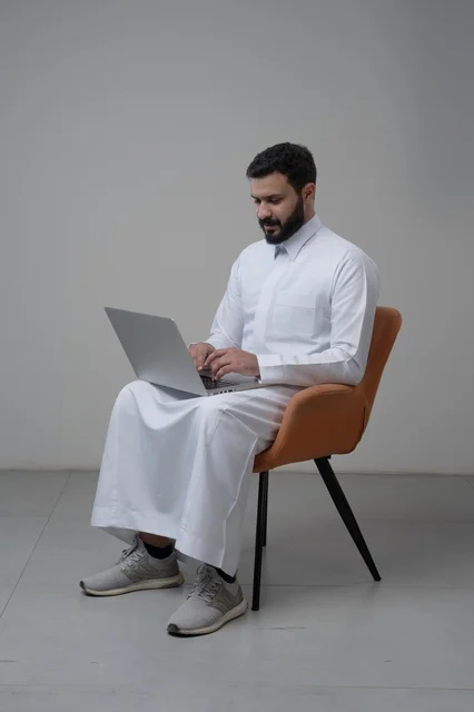 العمل عن بعد، استخدام التقنية الحديثه المتطورة في الحياة العملية، ايماءات الاندماج والتركيز، بورتريه لرجل عربي خليجي سعودي يرتدي الثوب التقليدي والشماغ يقوم بالعمل على الحاسب المحمول يجلس على كرسي مريح، خلفية بيضاء