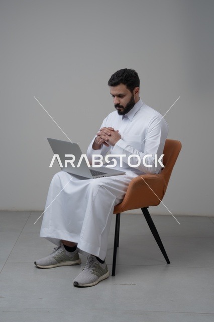 العمل عن بعد، استخدام التقنية الحديثه المتطورة في الحياة العملية، ايماءات الاندماج والتركيز، بورتريه لرجل عربي خليجي سعودي يرتدي الثوب التقليدي والشماغ يقوم بالعمل على الحاسب المحمول يجلس على كرسي مريح، خلفية بيضاء