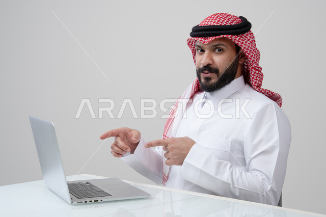  استخدام التقنية الحديثه المتطورة في الحياة العملية، بورتريه لرجل عربي خليجي سعودي مبتسم يرتدي الثوب التقليدي والشماغ يشير بيده الى الحاسب المحمول بتعابير وايماءات السرور والسعادة، مهنة ووظيفة ادارية، خلفية رمادية