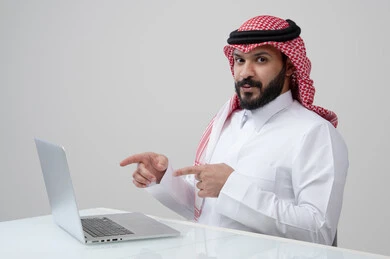  استخدام التقنية الحديثه المتطورة في الحياة العملية، بورتريه لرجل عربي خليجي سعودي مبتسم يرتدي الثوب التقليدي والشماغ يشير بيده الى الحاسب المحمول بتعابير وايماءات السرور والسعادة، مهنة ووظيفة ادارية، خلفية رمادية