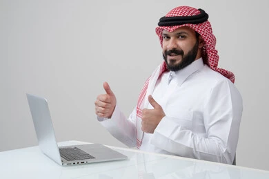 ايماءات الاعجاب والسرور، بورتريه لرجل عربي خليجي سعودي مبتسم يرتدي الثوب التقليدي والشماغ يرفع اصبع الابهام لأعلى بتعابير القبول يقوم بالعمل على الحاسب المحمول، مهنة ووظيفة ادارية، استخدام التقنية الحديثه المتطورة في الحياة العملية، خلفية رمادية