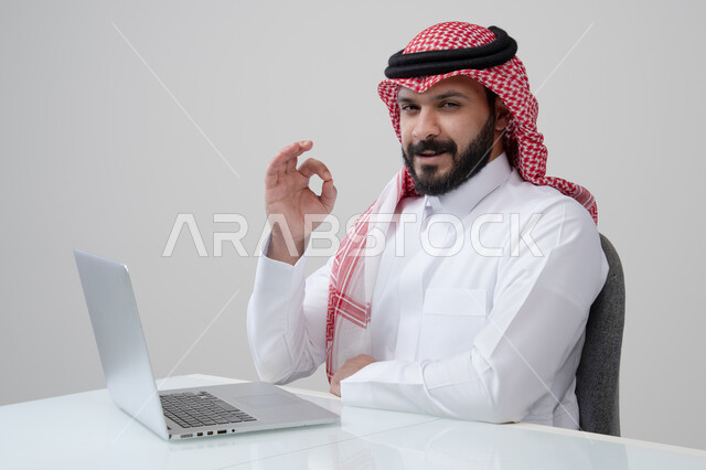 ايماءات التميز والامتياز، بورتريه لرجل عربي خليجي سعودي مبتسم يرتدي الثوب التقليدي والشماغ يرفع يده لأعلى بتعابير القبول يقوم بالعمل على الحاسب المحمول، مهنة ووظيفة ادارية، استخدام التقنية الحديثه المتطورة في الحياة العملية، خلفية رمادية