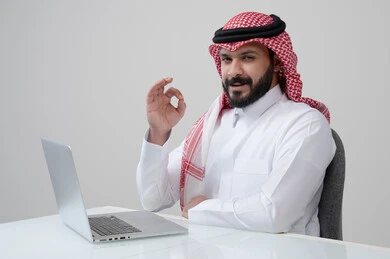 ايماءات التميز والامتياز، بورتريه لرجل عربي خليجي سعودي مبتسم يرتدي الثوب التقليدي والشماغ يرفع يده لأعلى بتعابير القبول يقوم بالعمل على الحاسب المحمول، مهنة ووظيفة ادارية، استخدام التقنية الحديثه المتطورة في الحياة العملية، خلفية رمادية