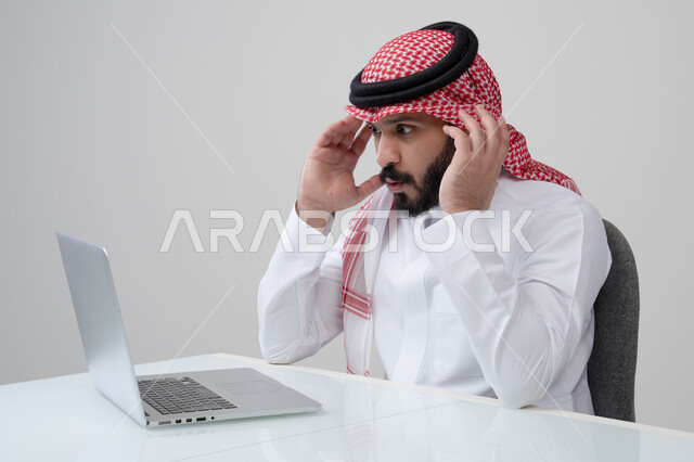 مفهوم ضغط العمل، مهنة ووظيفة ادارية، استخدام التقنية الحديثه المتطورة في الحياة العملية، ايماءات الارهاق والتعب، بورتريه لرجل عربي خليجي سعودي يرتدي الثوب التقليدي والشماغ يضع يده على رأسه بتعابير الصداع، العمل على الحاسب المحمول، خلفية رمادية