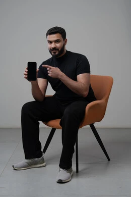 Pointing to a blank black screen, using modern advanced technology in practical life, working remotely via mobile, looking at the camera with gestures of pleasure, portrait of a young Saudi Arabian Gulf man wearing casual clothes sitting on a chair working on a mobile phone, gray background