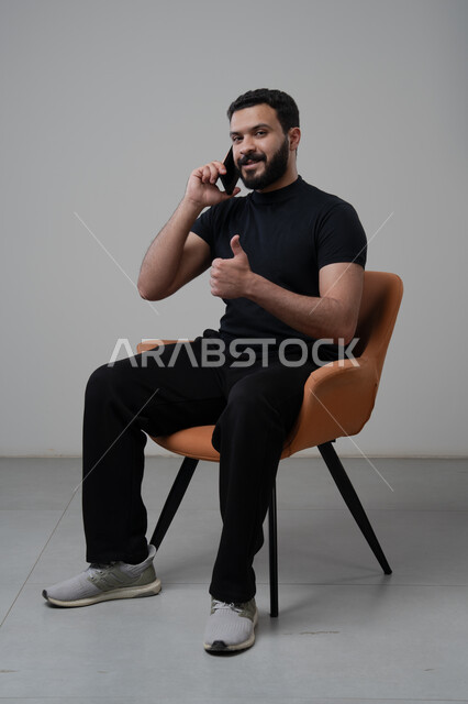رفع اصبع الابهام لأعلى بتعابير العجاب والقبول، الدردشة والمحادثة باستخدام جهاز تقني حديث, الرد على مكالمة هاتفية عبر الجوال، بورتريه لرجل عربي خليجي سعودي مبتسم يرتدي الزي الكاجوال يجلس على الكرسي يقوم باجراء اتصال هاتفي من خلال الهاتف المحمول، خلفية رمادية