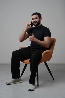 رفع اصبع الابهام لأعلى بتعابير العجاب والقبول، الدردشة والمحادثة باستخدام جهاز تقني حديث, الرد على مكالمة هاتفية عبر الجوال، بورتريه لرجل عربي خليجي سعودي مبتسم يرتدي الزي الكاجوال يجلس على الكرسي يقوم باجراء اتصال هاتفي من خلال الهاتف المحمول، خلفية رمادية