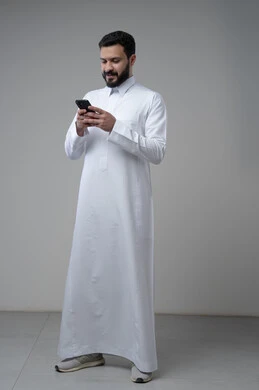 Gestures and expressions of integration and concentration while working on the mobile phone, use of technology and technology in daily life, portrait of a Saudi Arabian Gulf man wearing traditional thobe holding a mobile phone in his hand, full body image, gray background