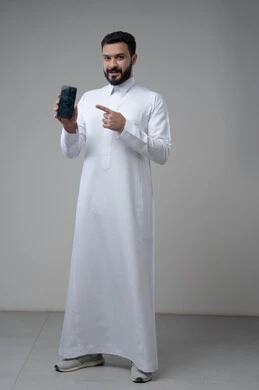 عرض شاشة سوداء فارغة عبر الجوال، استخدام التقنية والتكنولوجيا في الحياة اليومية، بورتريه لرجل عربي خليجي سعودي يرتدي الثوب التقليدي يقوم بالاشارة الى الهاتف المحمول، صورة الجسم بالطول الكامل، خلفية رمادية
