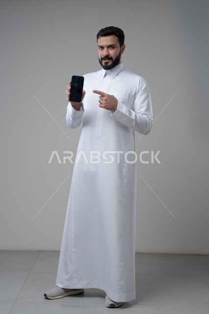 عرض شاشة سوداء فارغة عبر الجوال، استخدام التقنية والتكنولوجيا في الحياة اليومية، بورتريه لرجل عربي خليجي سعودي يرتدي الثوب التقليدي يقوم بالاشارة الى الهاتف المحمول، صورة الجسم بالطول الكامل، خلفية رمادية