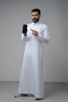 عرض شاشة سوداء فارغة عبر الجوال، استخدام التقنية والتكنولوجيا في الحياة اليومية، بورتريه لرجل عربي خليجي سعودي يرتدي الثوب التقليدي يقوم بالاشارة الى الهاتف المحمول، صورة الجسم بالطول الكامل، خلفية رمادية