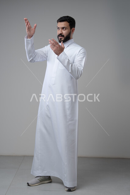 النظر الى الاعلى بتعابير التركيز، بورتريه لرجل عربي خليجي سعودي يرتدي الثوب التقليدي يشير بيده الى شيء ما، الدلالة على مكان معين، صورة الجسم بالطول الكامل، خلفية رمادية