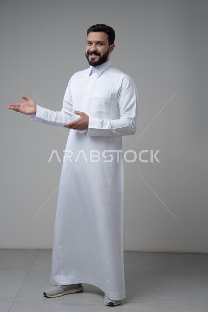 الترحيب والاستقبال بتعابير السرور والبهجة، بورتريه لرجل عربي خليجي سعودي مبتسم يرتدي الثوب التقليدي يشير بيده الى مكان ما، الدلالة على مكان معين، صورة الجسم بالطول الكامل، خلفية رمادية