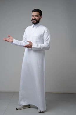 Welcoming and receiving with expressions of pleasure and joy, portrait of a smiling Saudi Arabian Gulf man wearing traditional thobe pointing to a place, indicating a specific place, full-length body image, gray background