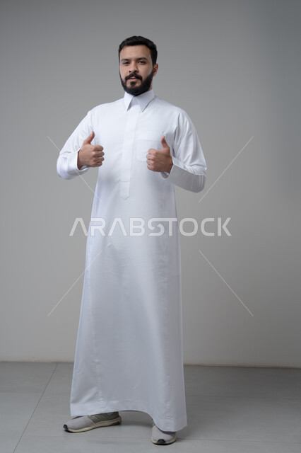 رفع اصابع الابهام الى الاعلى، النظر الى الكاميرا  بتعابير الاعجاب والرضا، بورتريه لرجل عربي خليجي سعودي يرتدي الثوب التقليدي ينظر الى الكاميرا بايماءات القبول والموافقة، صورة الجسم بالطول الكامل، خلفية رمادية