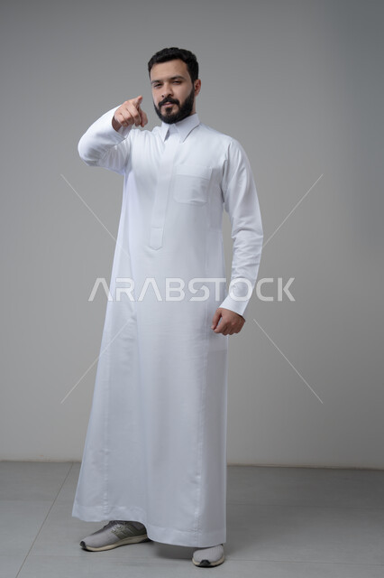 Pointing forward, looking at the camera with a serious and firm expression, portrait of a Saudi Arabian Gulf man wearing traditional thobe pointing at something, indicating a specific place, full body image, gray background