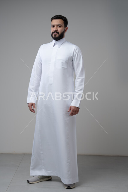  الاعتناء والاهتمام بالمظهر الخارجي، الوقوف بجدية وحزم، مفهوم الاناقة والرجولة، بورتريه لشاب عربي خليجي سعودي يرتدي الثوب التقليدي يقف باستقامة ينظر الى الكاميرا بتعابير الثقة بالنفس، الجسم بالطول الكامل، خلفية رمادية